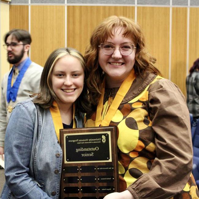 two graduating seniors smile with a plaque